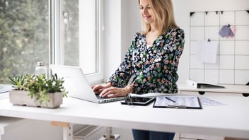 Smiling woman typing on a laptop at a bright desk beside a window