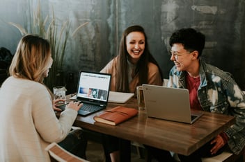 Three coworkers laughing and collaborating around a table with laptops