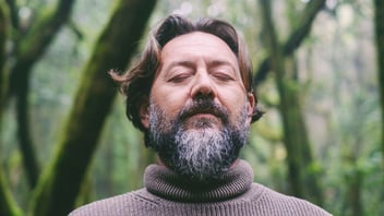 Bearded man standing among green forest trees with his eyes closed in a calm moment