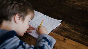 Boy working through math homework in a notebook at a wooden kitchen table