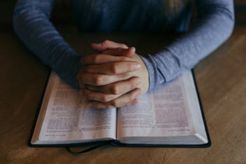 Hands folded in prayer resting on an open Bible on a wooden table
