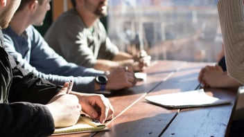 Colleagues sit around a sunlit wooden table with notepads during a team meeting