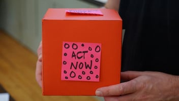 Hands holding a red donation box with a handwritten act now note