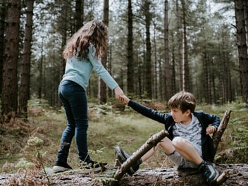 Older sister helping her younger brother climb over a log in the woods