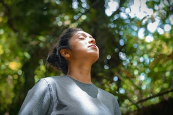 Woman with eyes closed taking a calming breath outdoors under sunlit trees