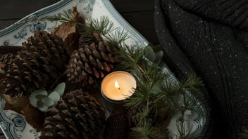 A lit tealight candle rests among pinecones and evergreen sprigs beside a dark knit blanket