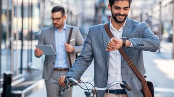 A smiling man wheeling a bicycle checks his watch on a city sidewalk while a colleague walks behind him