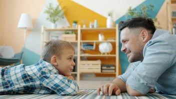 A father and young son lie on the living room floor, talking face to face