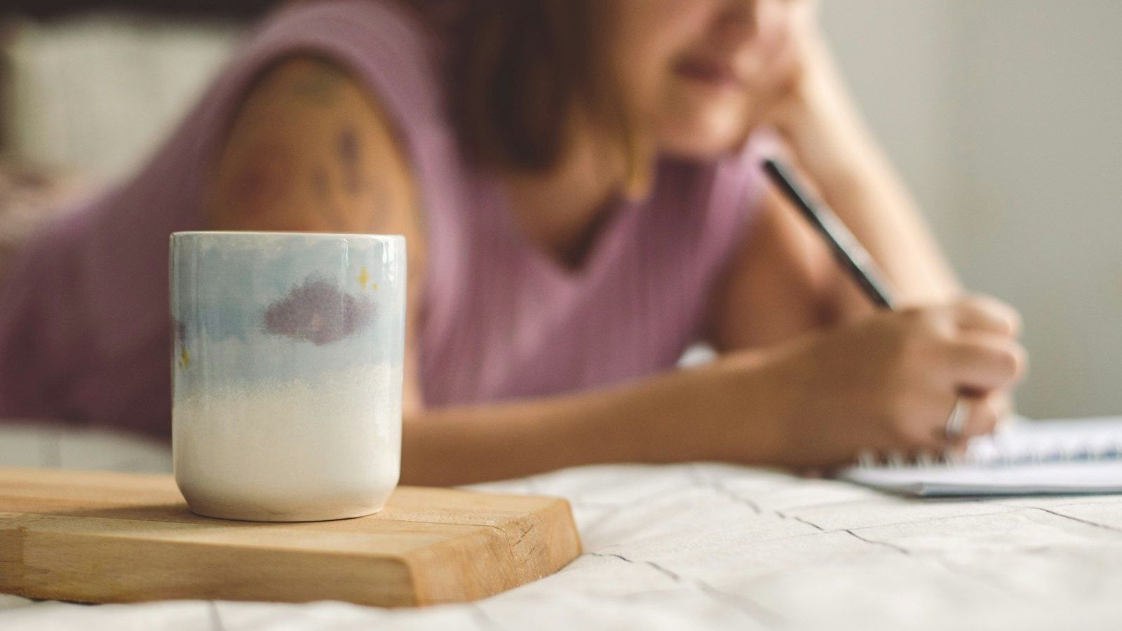 A woman writes in a notebook beside a hand-painted mug in soft morning light