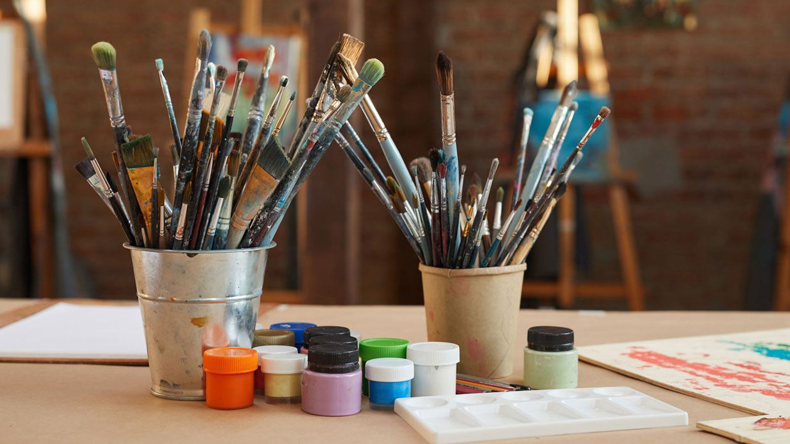 Paintbrushes standing in two cups beside colorful paint pots on an art studio table