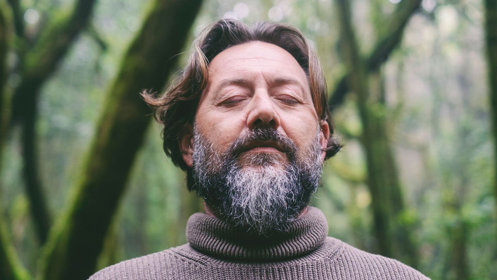Bearded man standing among green forest trees with his eyes closed in a calm moment