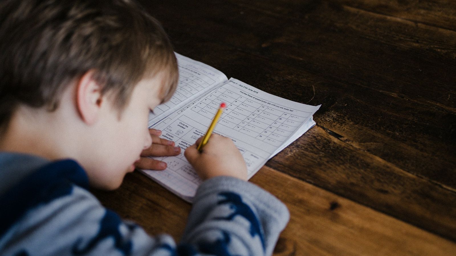 Boy working through math homework in a notebook at a wooden kitchen table
