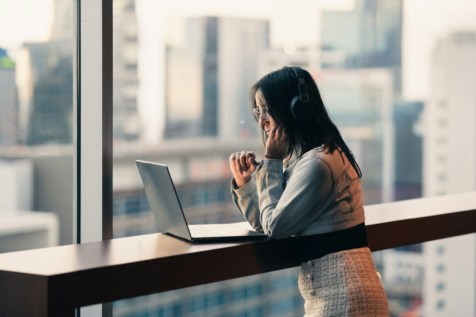 Professional woman with headphones resting her chin on her hand at a laptop by a city window at the end of the day