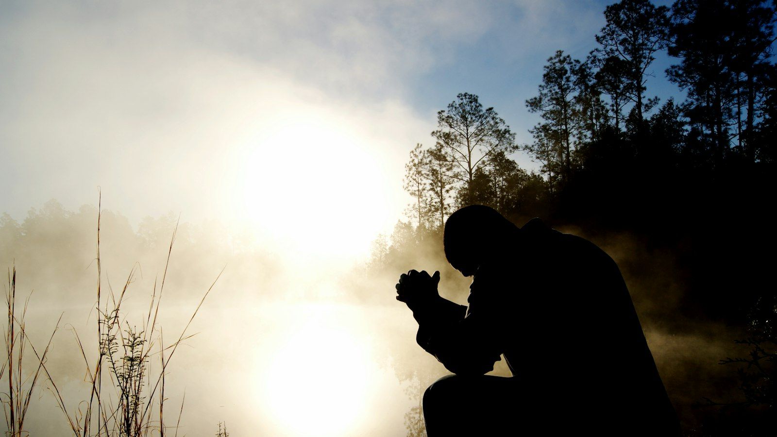 Silhouette of a man kneeling in prayer beside a misty lake at sunrise