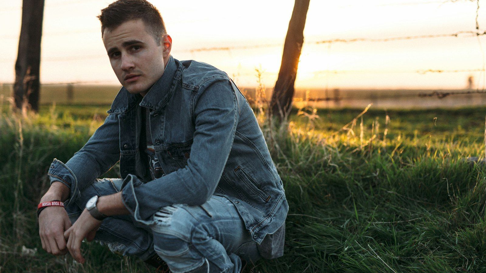 A pensive young person crouches in a grassy field at sunset