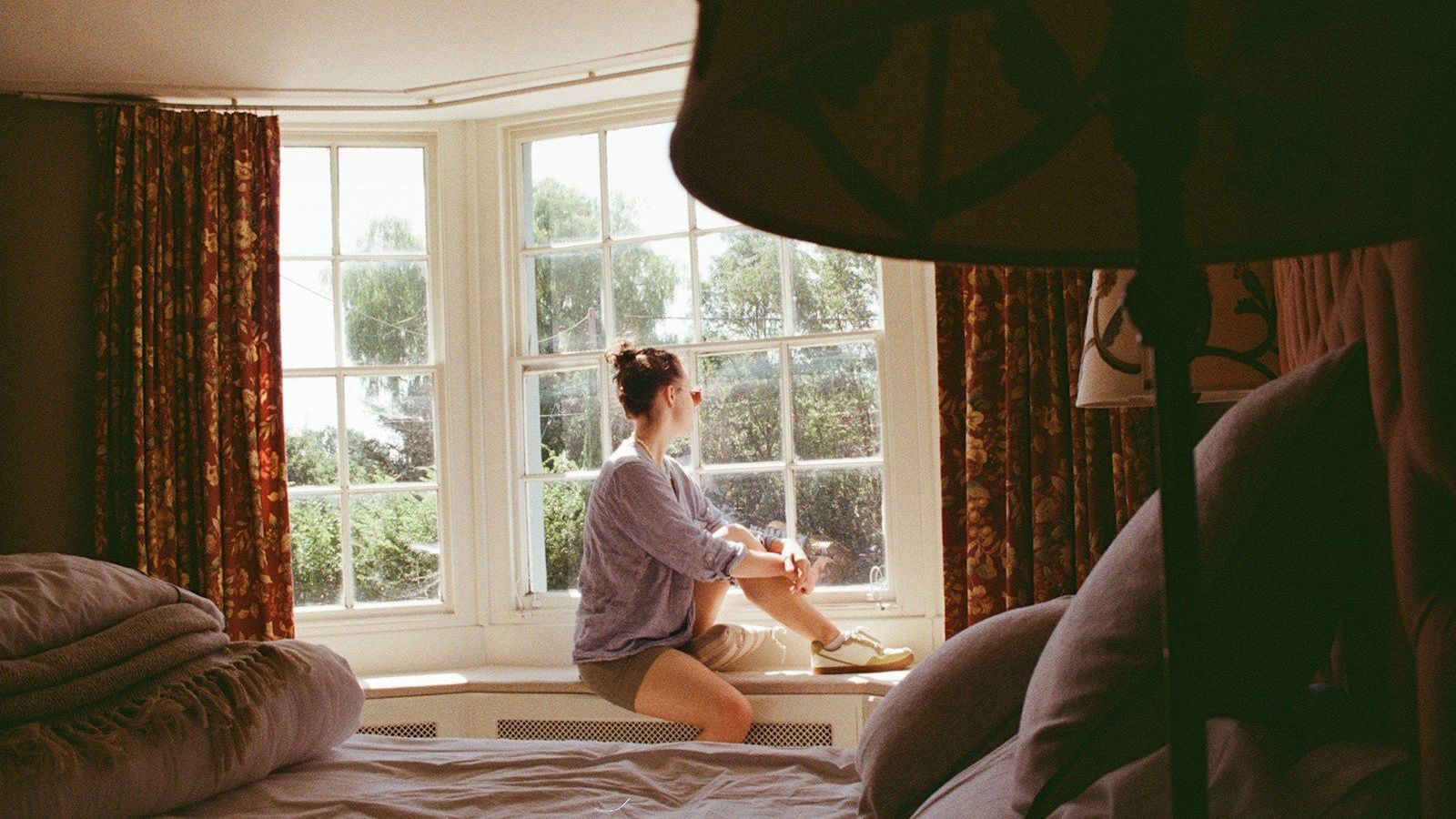 A woman sits on a sunlit window seat, gazing out at the trees