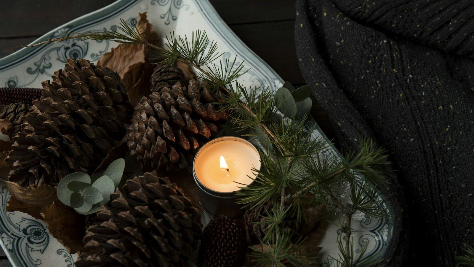 A lit tealight candle rests among pinecones and evergreen sprigs beside a dark knit blanket