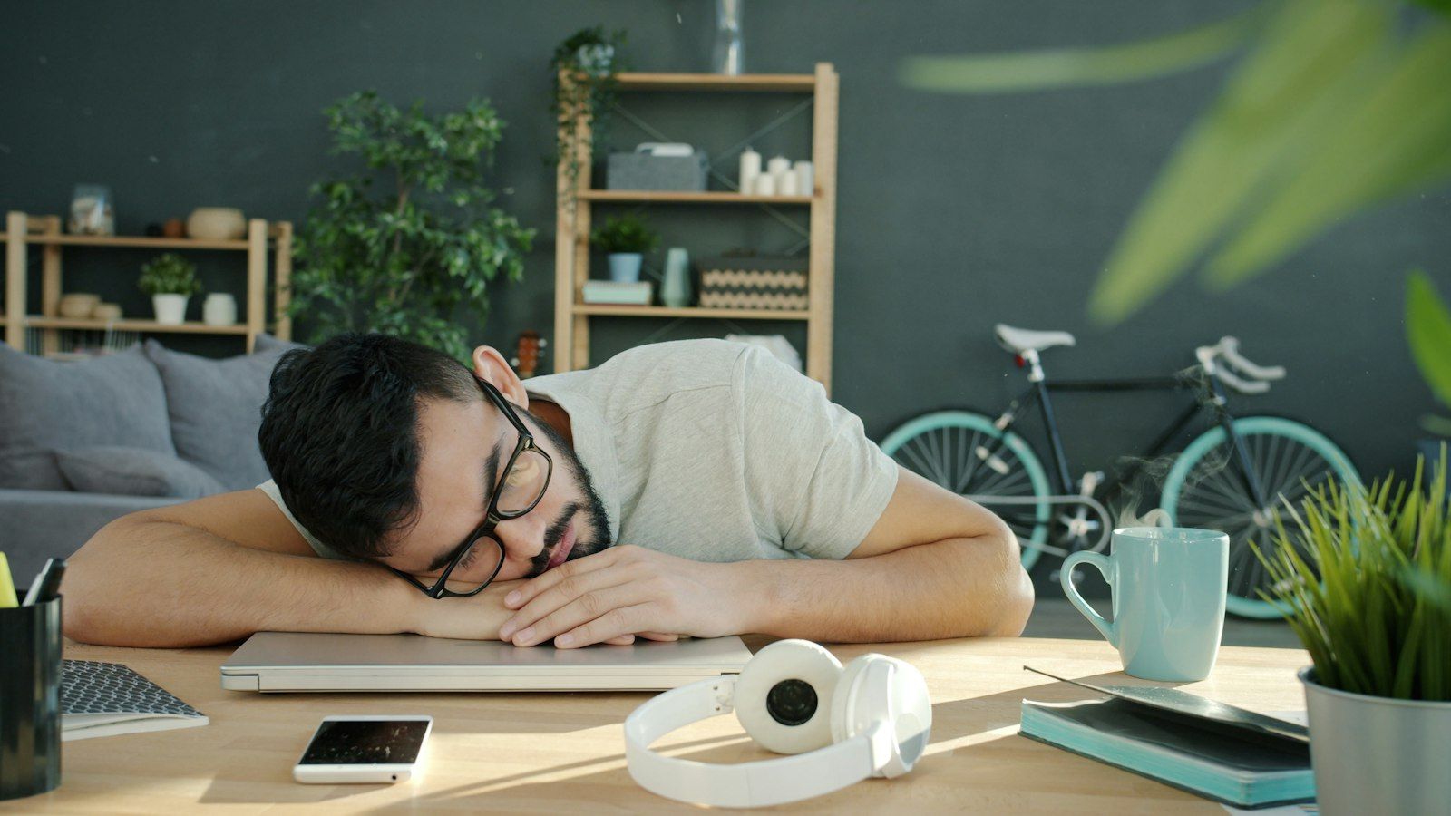 A man dozes on his laptop at a desk beside a steaming cup of coffee