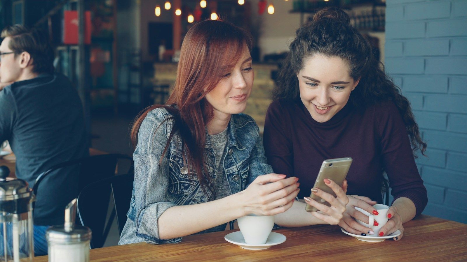Two friends smiling at a smartphone together in a cafe