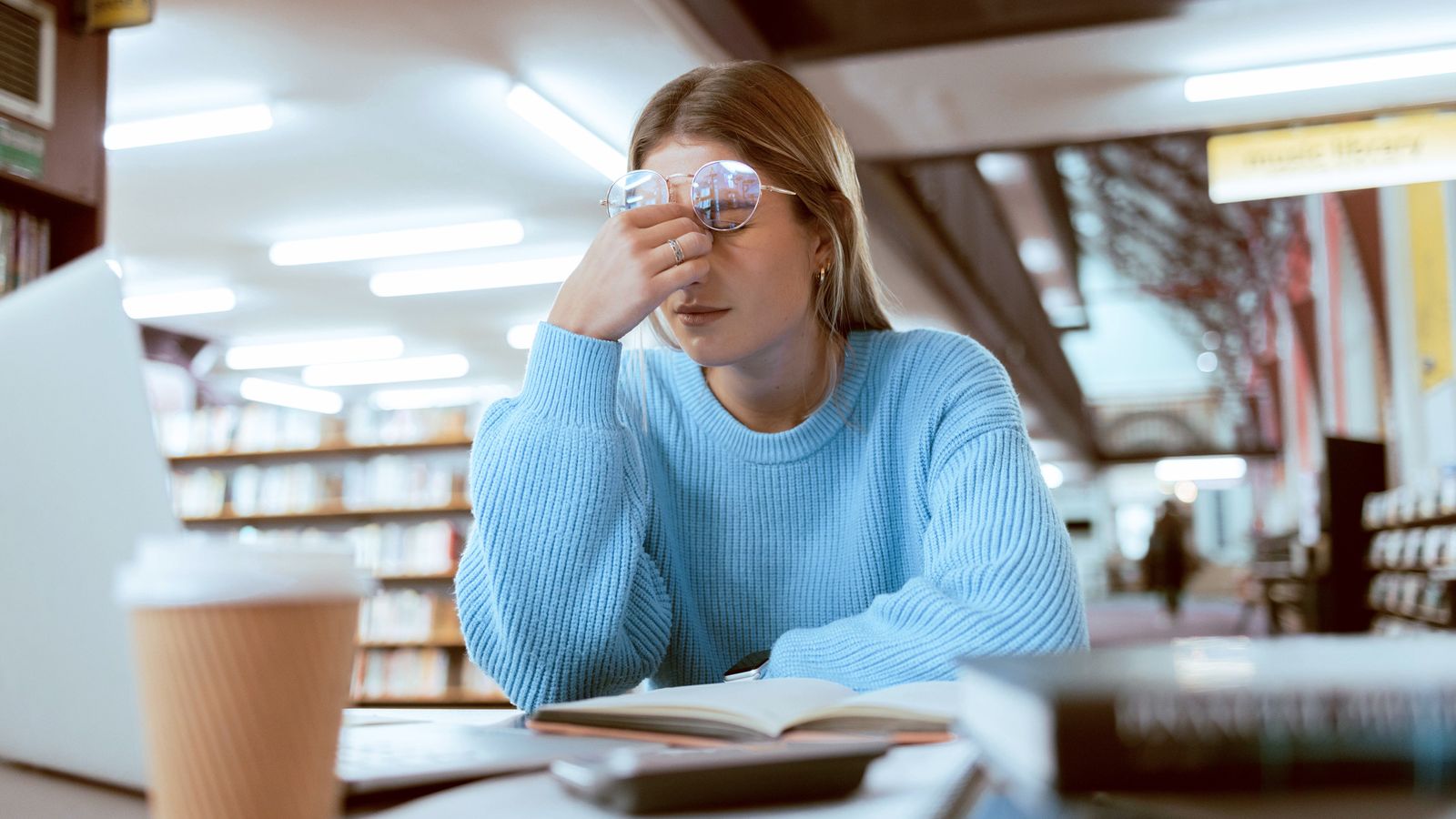 Young woman pausing over a book in a library, rubbing her tired eyes