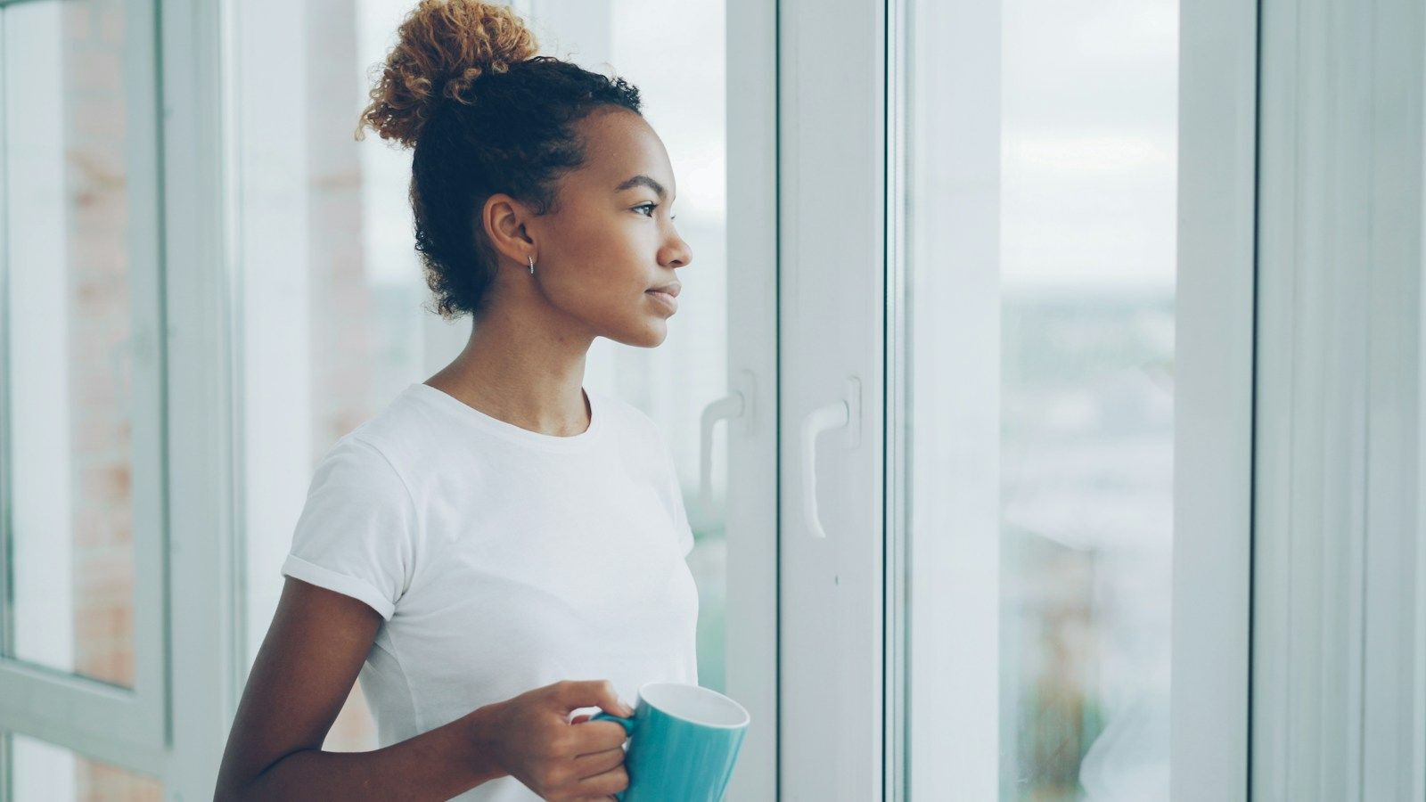 Woman holding a mug and looking thoughtfully out a bright window