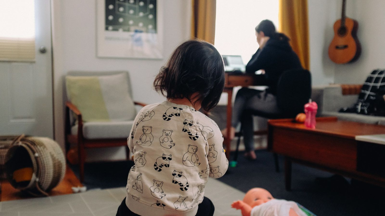 Toddler playing in the living room while their mother works at a laptop in the background
