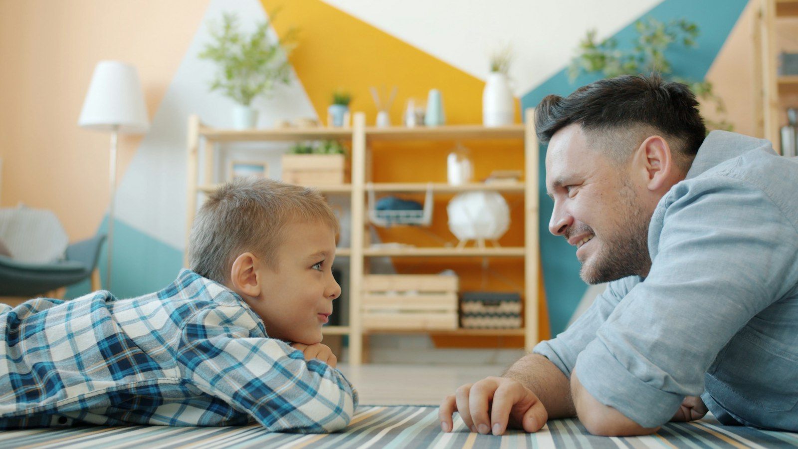 A father and young son lie on the living room floor, talking face to face
