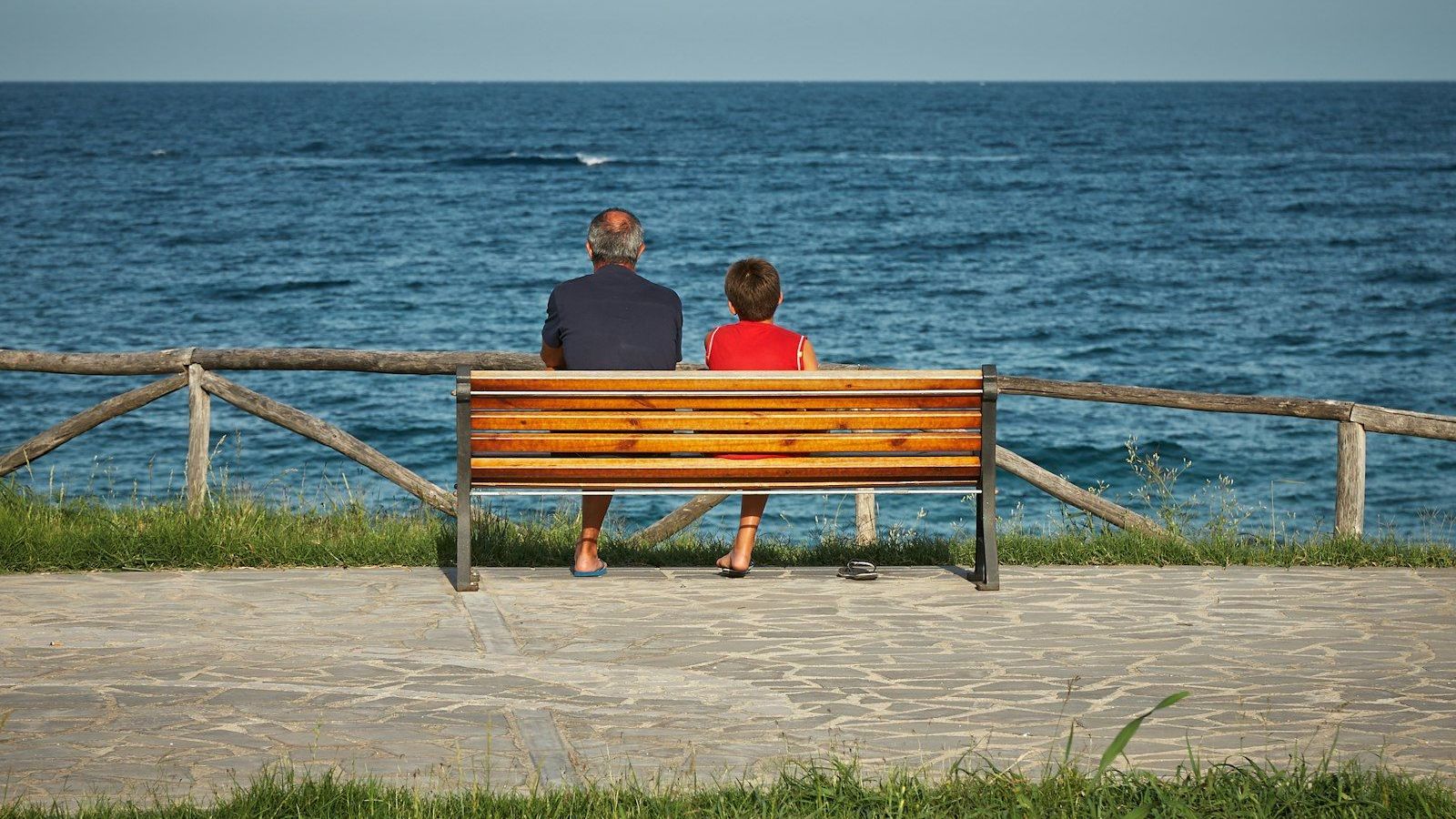 An adult and a child sit together on a bench overlooking the ocean