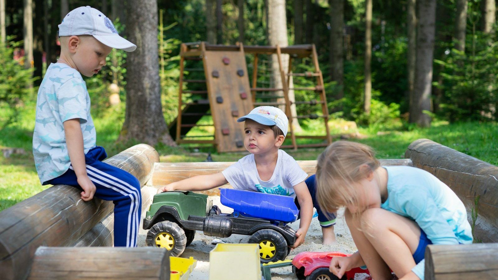 Three young children play together with toy trucks in a sandbox