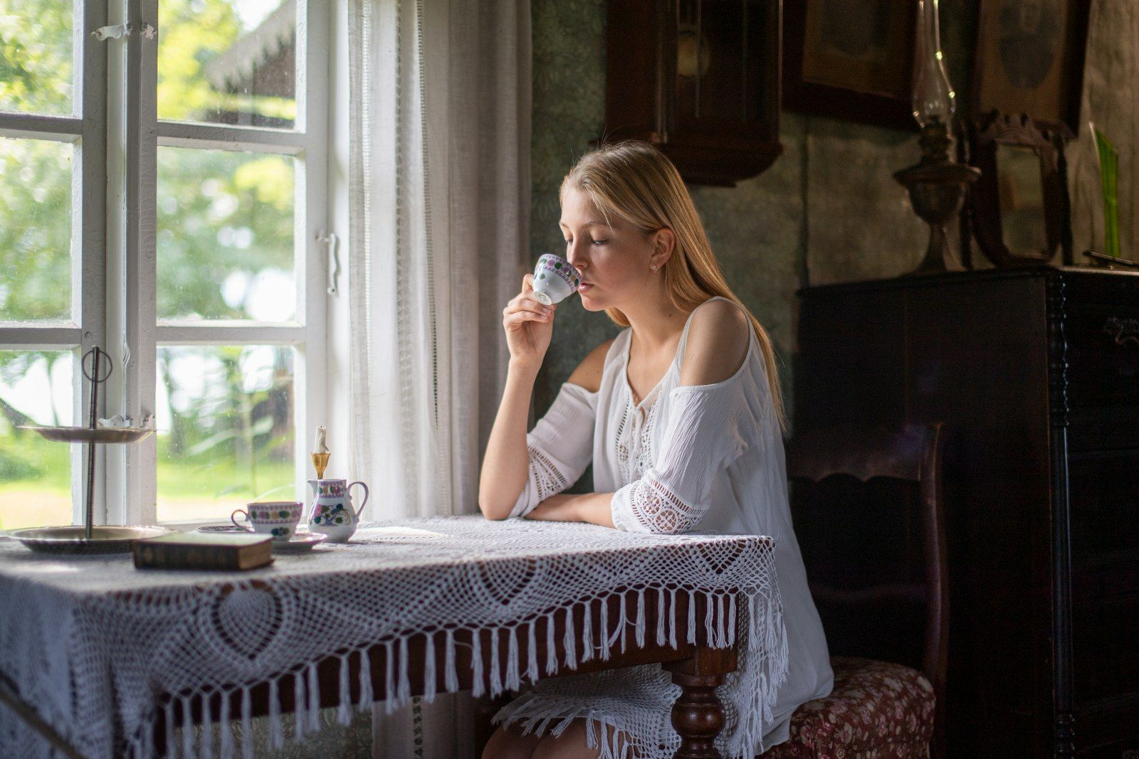 A woman sips tea calmly at a lace covered table beside a bright cottage window