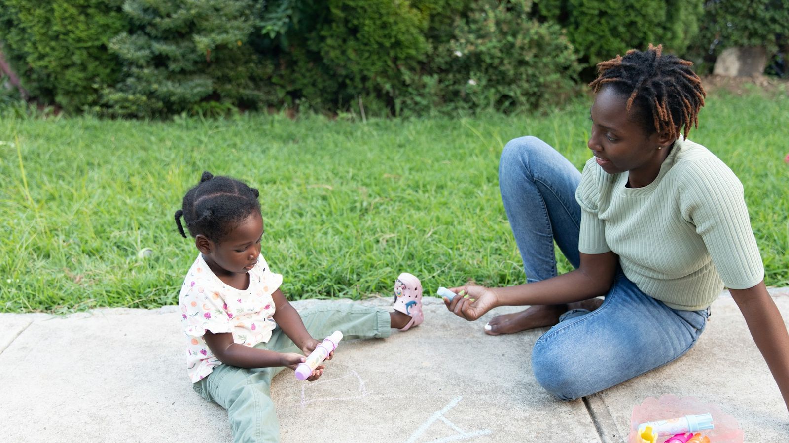 Parent sitting on the sidewalk drawing with chalk alongside her young child