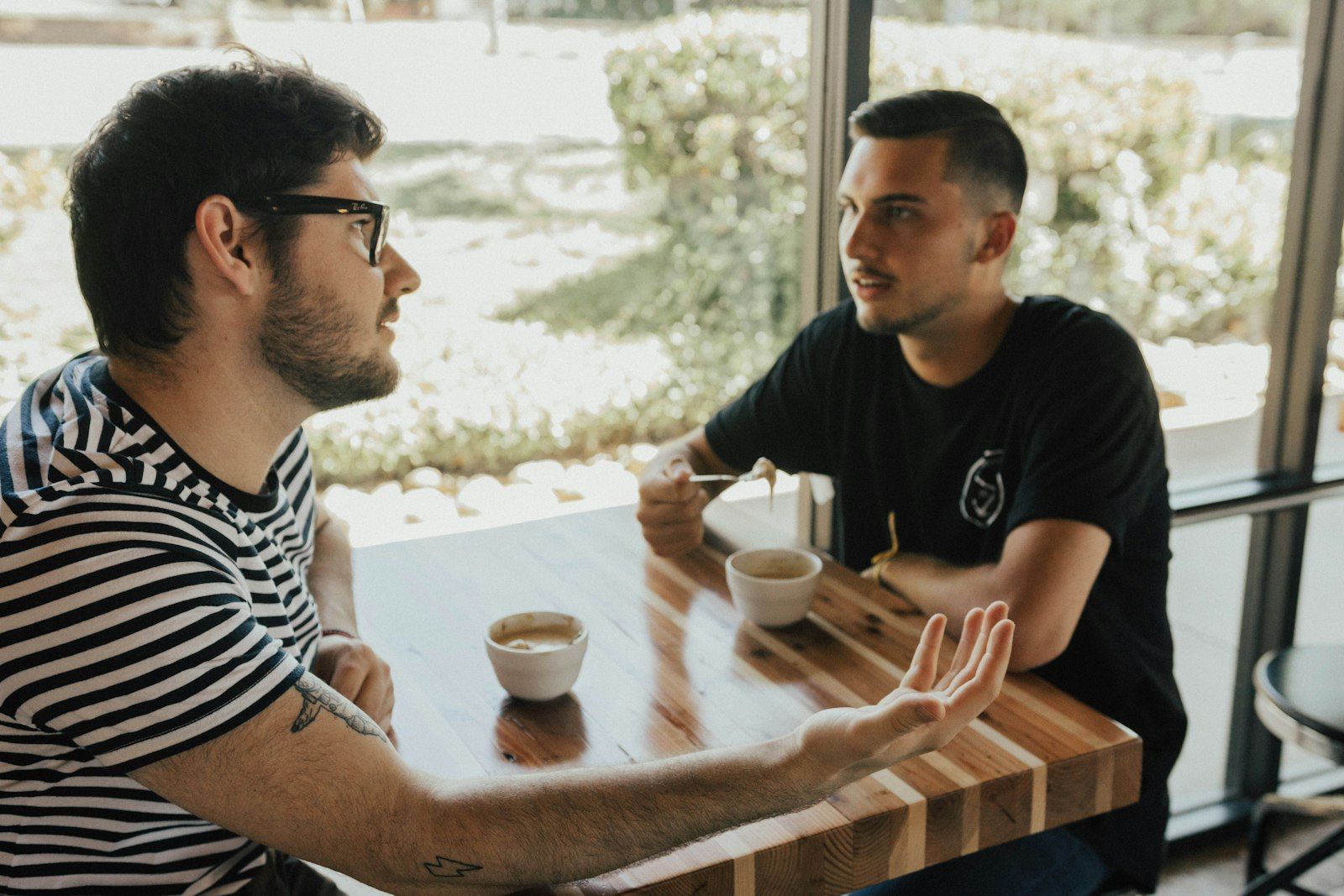 Man gesturing with an open hand as he talks with another man over coffee at a sunlit table